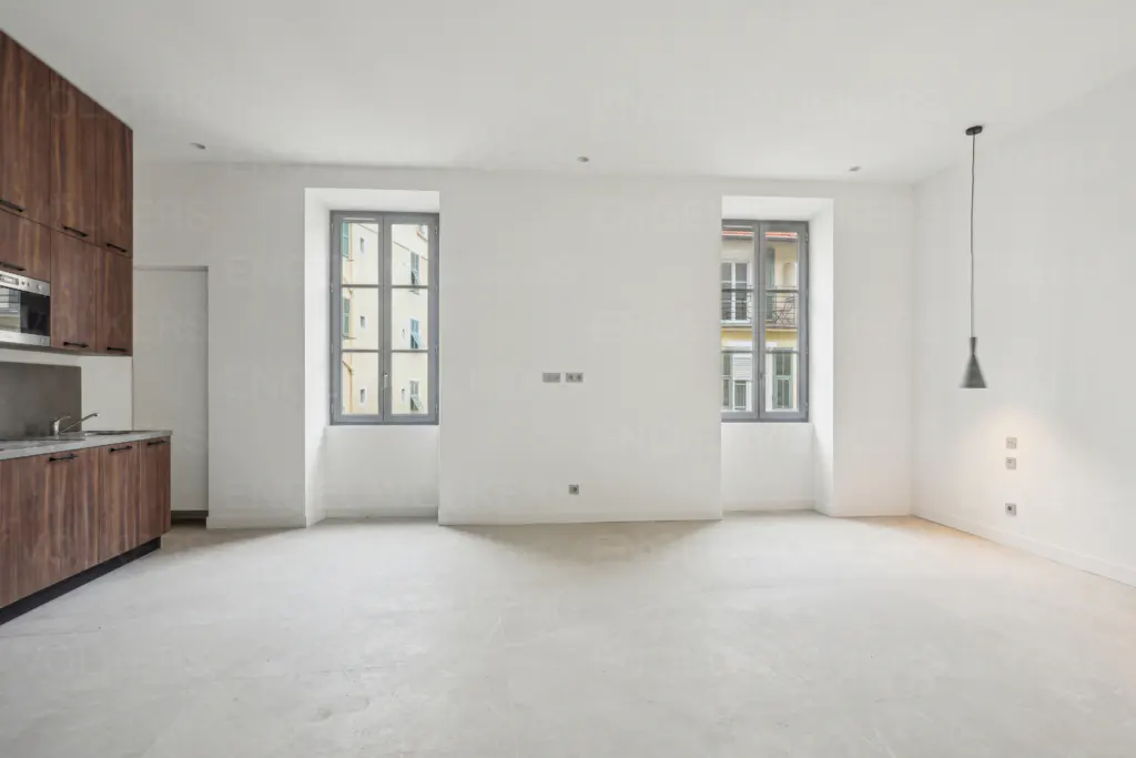 Bright, empty apartment with white walls, gray windows, and wood cabinets. A pendant light hangs in the corner.