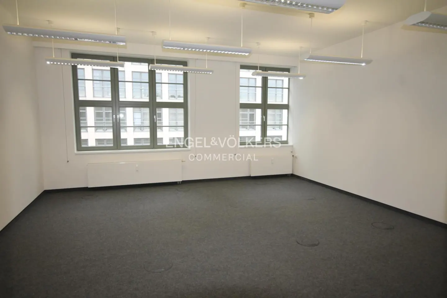 Empty office space with gray carpet, white walls, and two windows with green frames. Overhead lighting and radiators are visible.