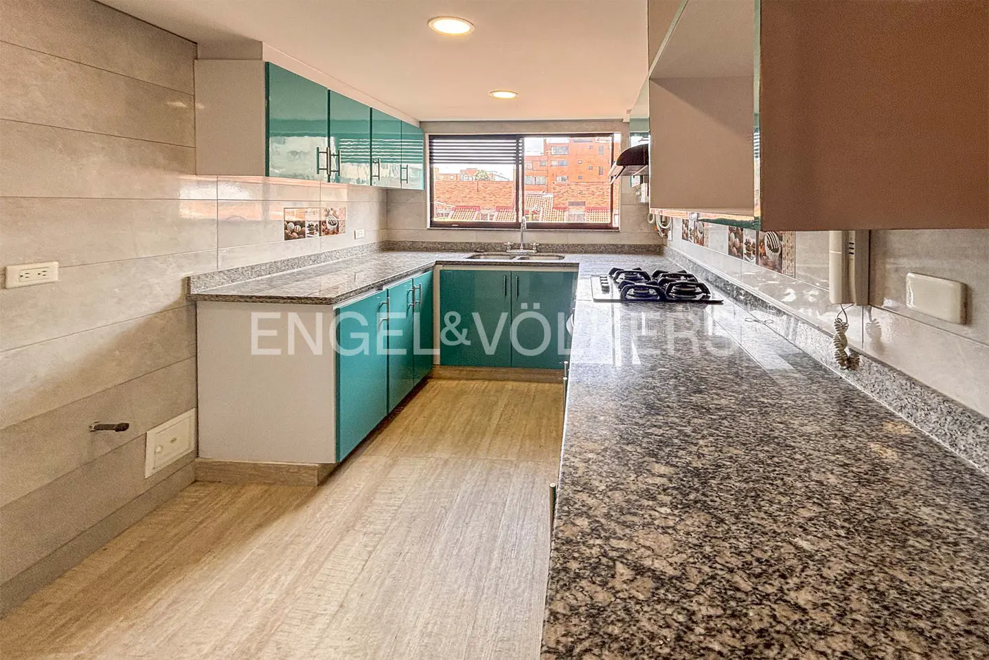 A modern kitchen with teal cabinets, granite countertops, and a gas stovetop. A window overlooks a cityscape.