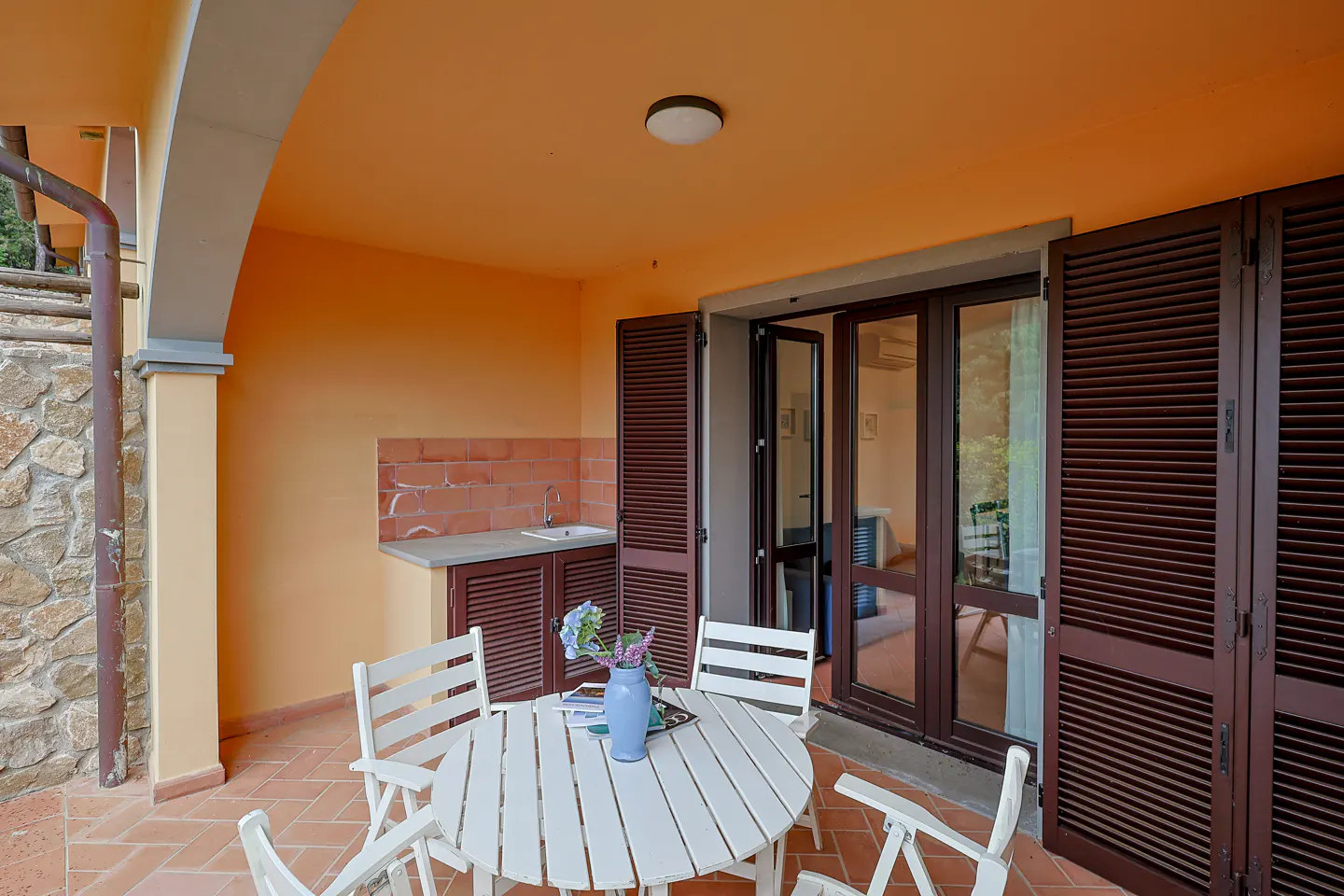 Outdoor patio with white table, chairs, and blue vase. Orange walls, brown shutters, and a small outdoor kitchen area.