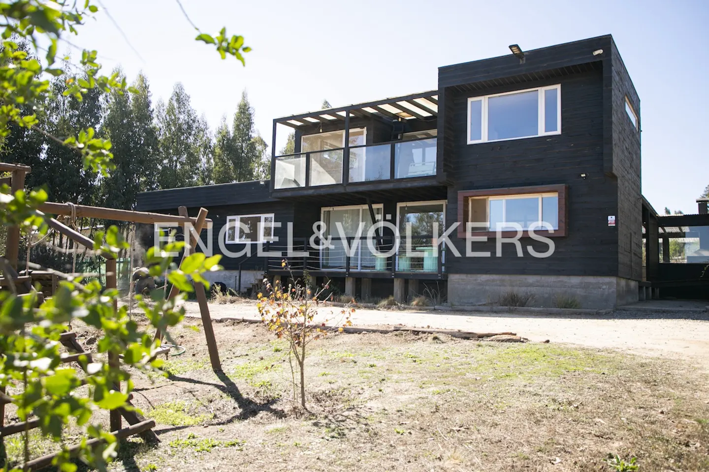 Two-story modern house with black wood siding, large windows, and a balcony with glass railings. A wooden swing set is in the yard.