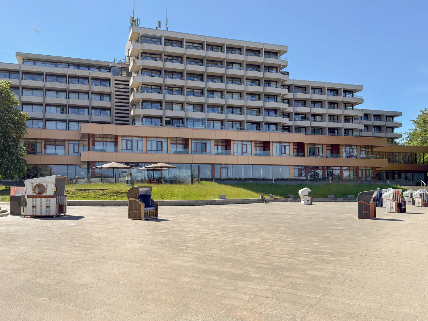 Large multi-story hotel with balconies overlooking a paved plaza featuring several traditional hooded beach chairs.