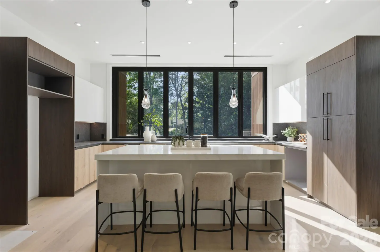 Modern kitchen with a white island, four beige bar stools, dark wood cabinetry, and a large window overlooking trees.