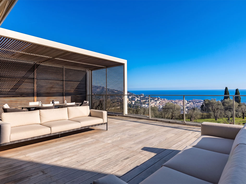 Spacious wooden terrace with beige sofas under a slatted pergola, overlooking a coastal city and the blue sea. [AI-generated text]