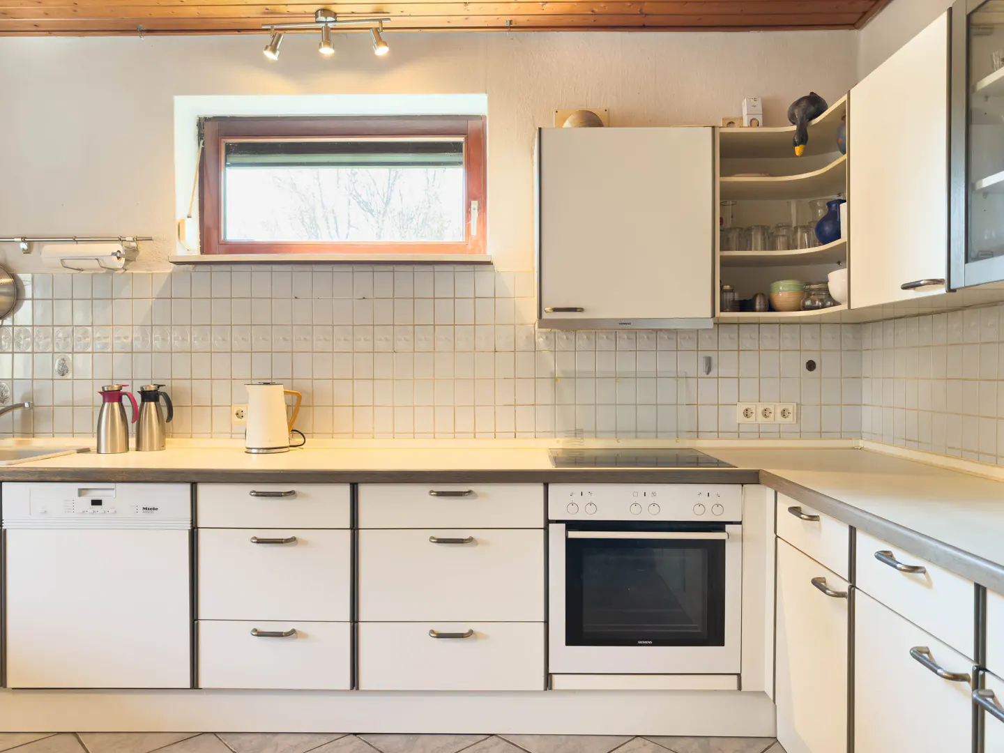 Bright kitchen with white cabinets, tile backsplash, and wood trim window. Appliances include a dishwasher, oven, and electric kettle.