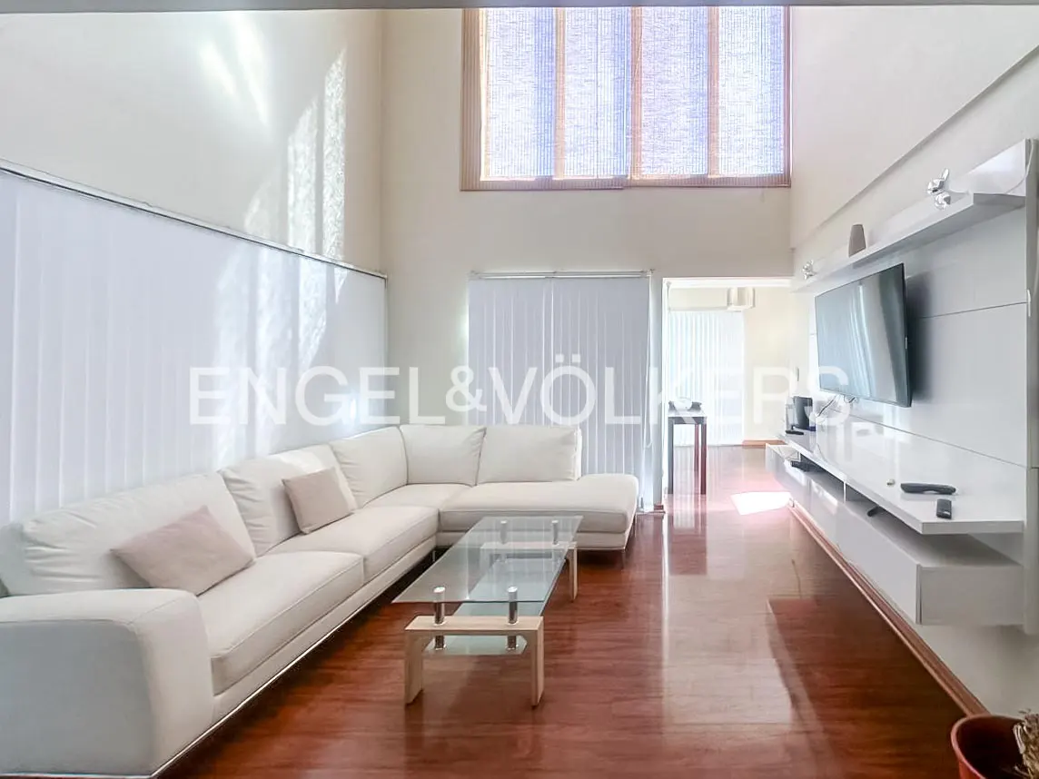 Bright living room with white sectional sofa, glass coffee table, and wood floors. A large TV is mounted on a white wall unit.