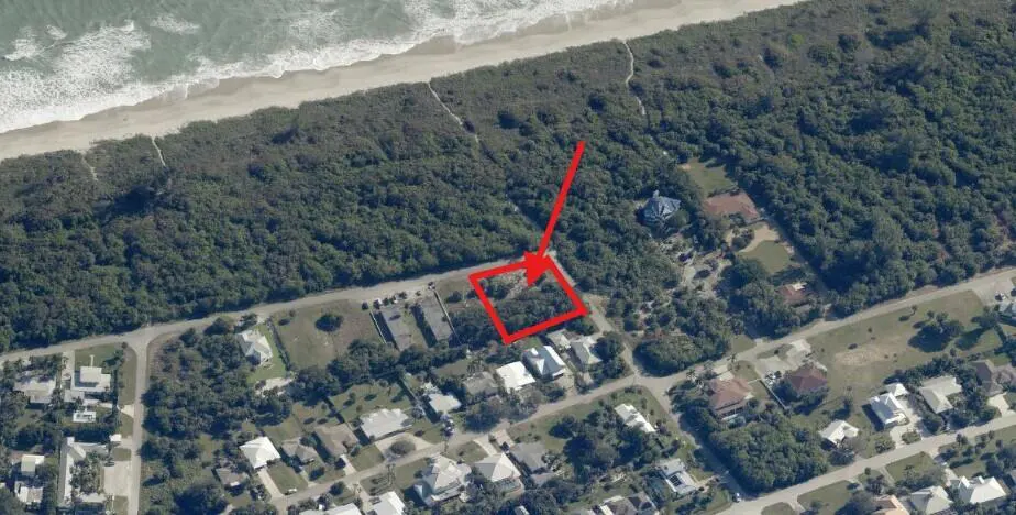 Aerial view of a property near the beach, marked with a red square and arrow, surrounded by trees and houses.