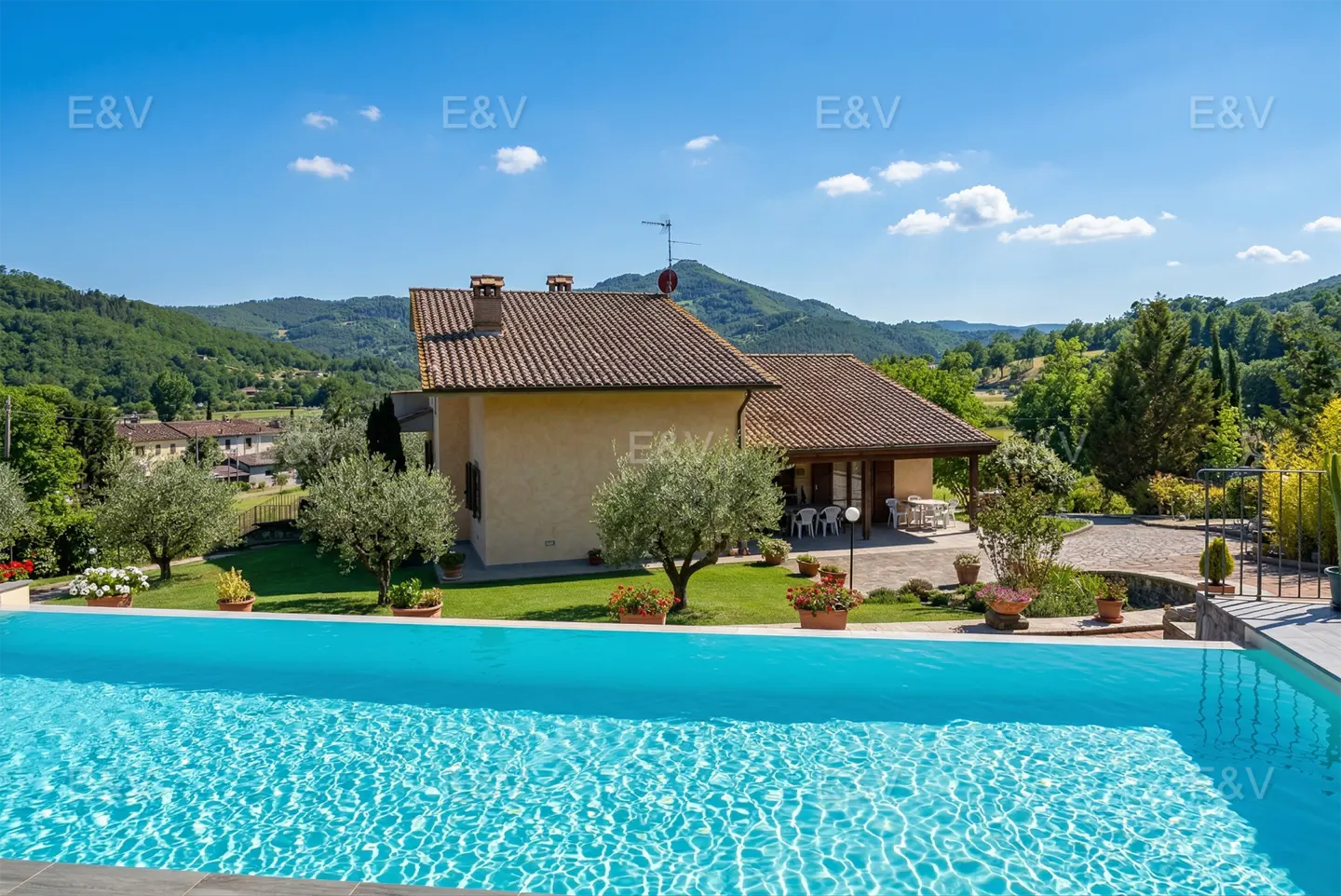 A tan house with a brown roof overlooks a blue swimming pool on a sunny day. Green hills are in the background.