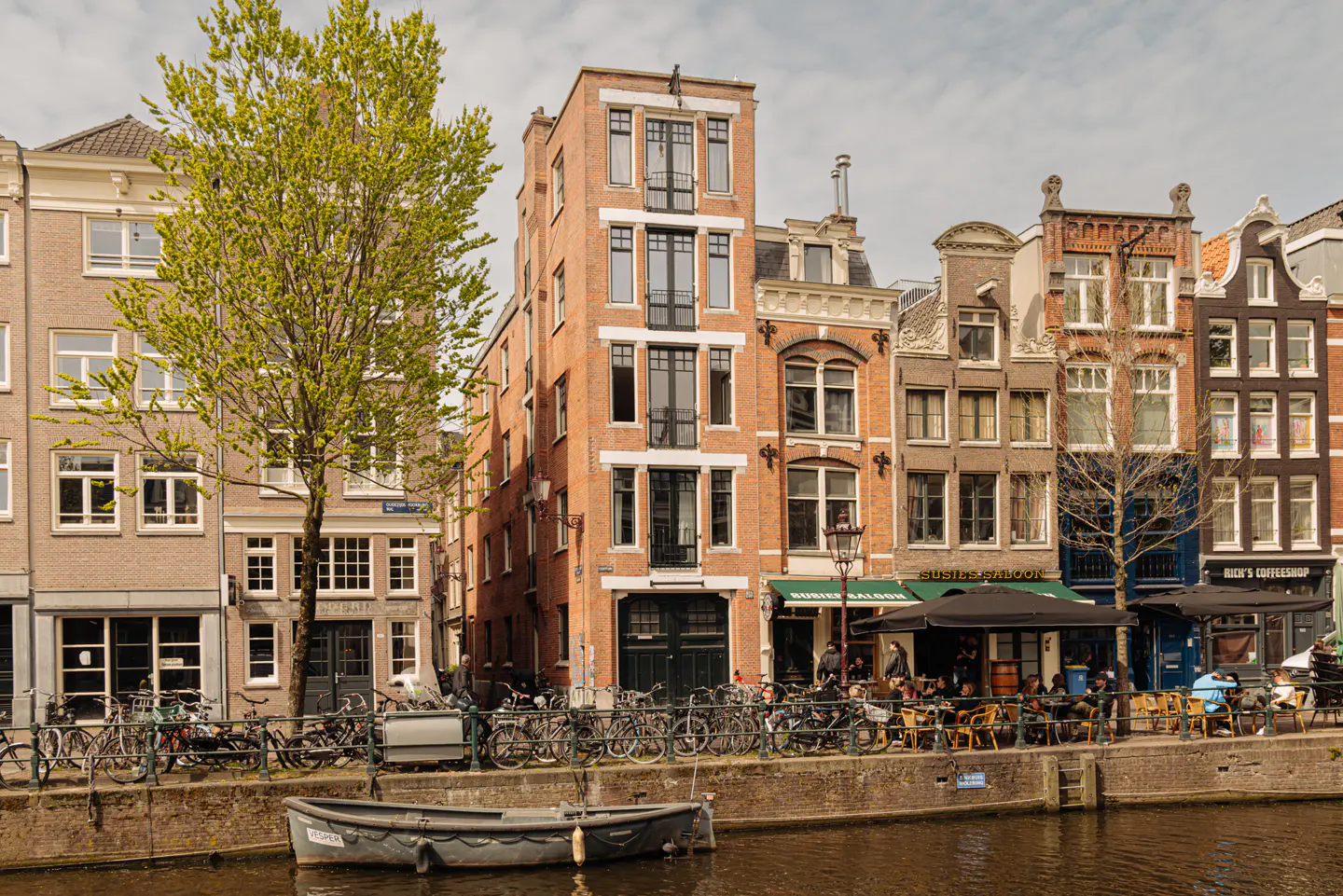 Amsterdam canal view with brick buildings, cafes, and bicycles. A small boat floats on the water.
