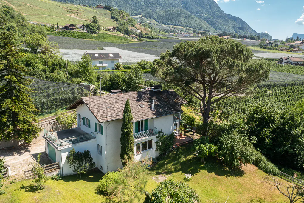 A white two-story house with a brown roof sits in a lush green valley surrounded by vineyards and mountains.