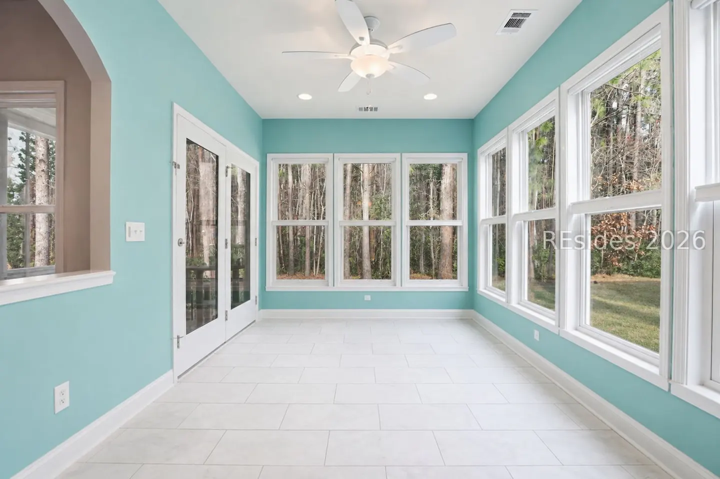 Bright sunroom with white tile floor, aqua walls, and white trim. Large windows offer a view of trees. A white ceiling fan is centered.