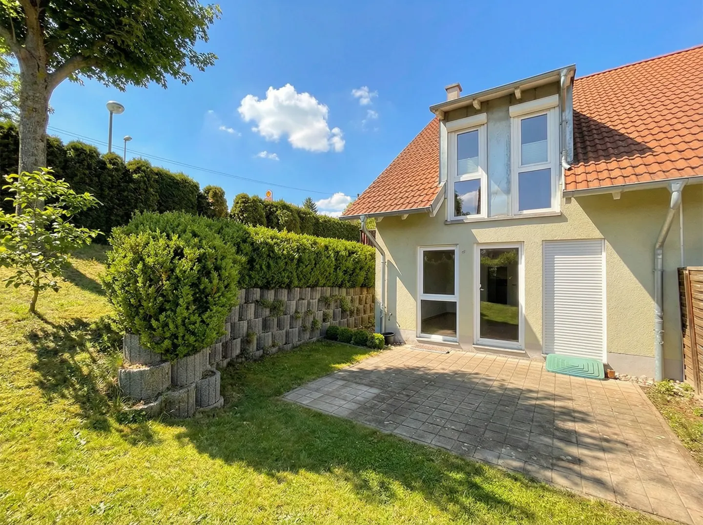 Backyard view of a two-story house with a red tile roof, a stone patio, and a green lawn. A tall hedge provides privacy.