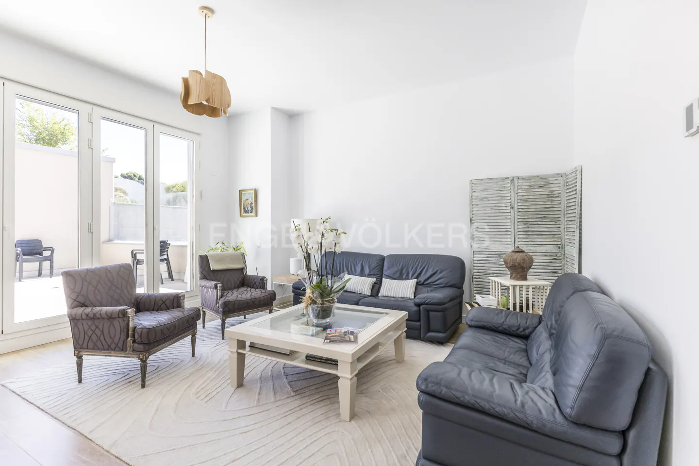 Bright living room with two blue leather sofas, two patterned armchairs, and a white coffee table on a patterned rug. Patio doors lead to an outdoor seating area.