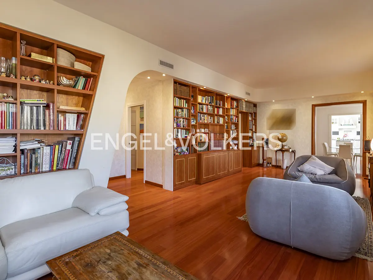 A spacious living room with hardwood floors, bookshelves, and comfortable seating. A white sofa and gray chairs are visible.