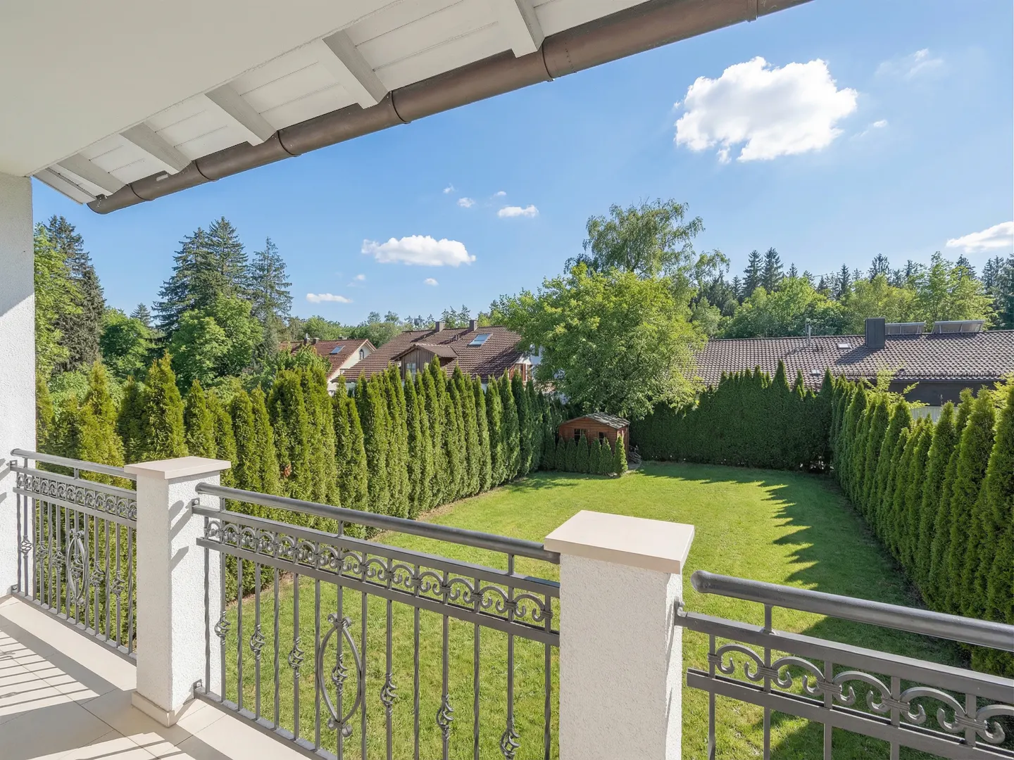 Balcony view of a green lawn surrounded by tall hedges, with houses and trees in the background under a blue sky.