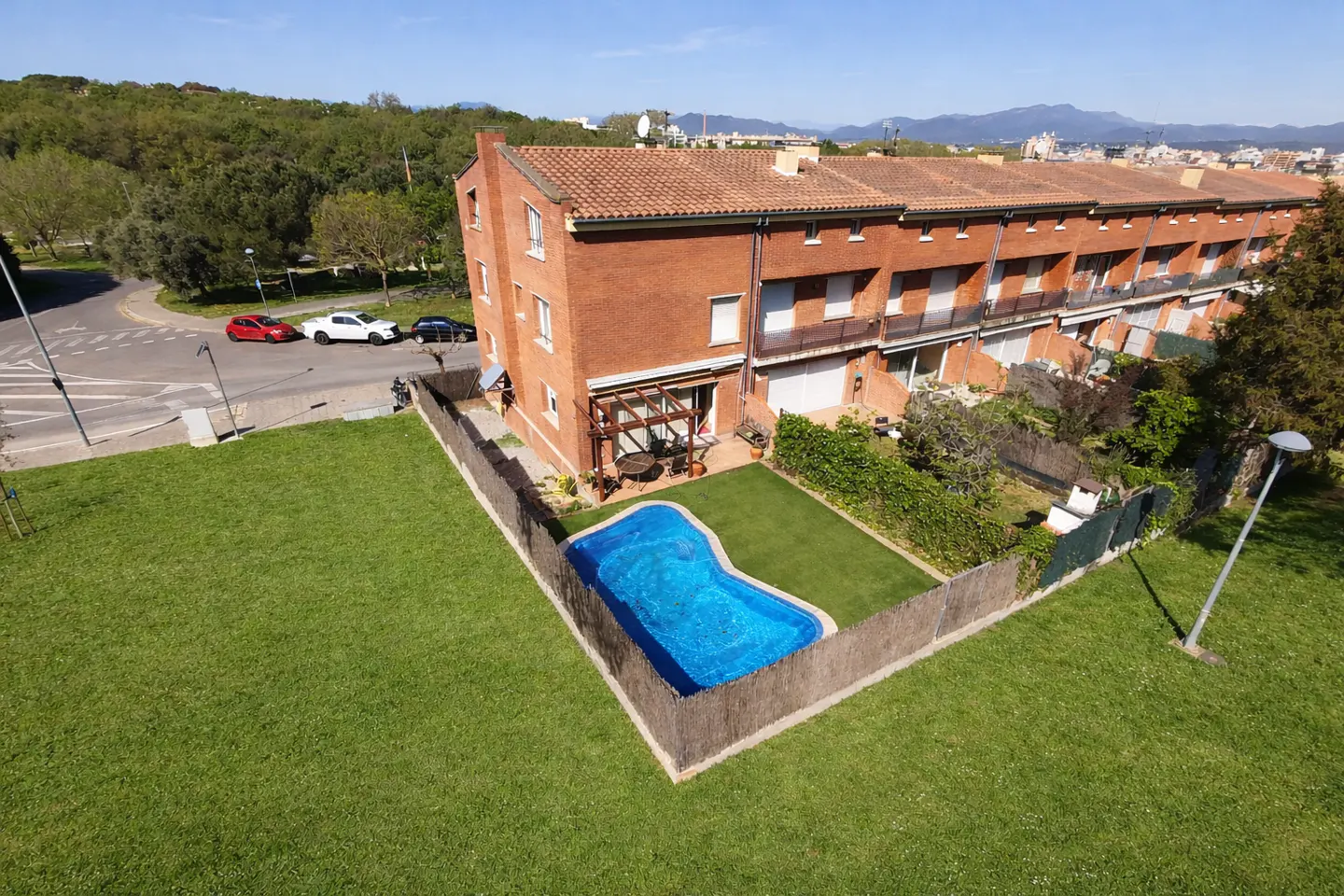 Aerial view of brick townhouses with a blue pool in a fenced backyard, green lawn, and cars parked on the street.
