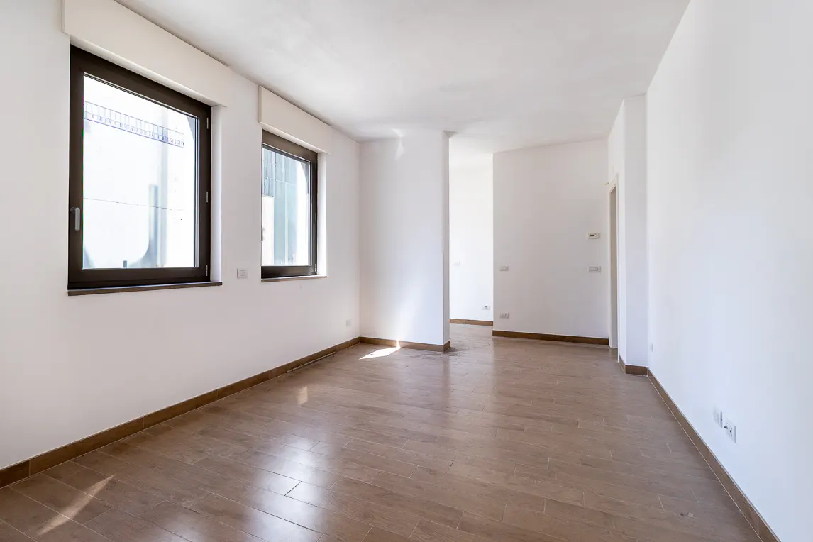 Bright, empty room with white walls, brown wood-look tile floor, and two dark-framed windows letting in natural light.