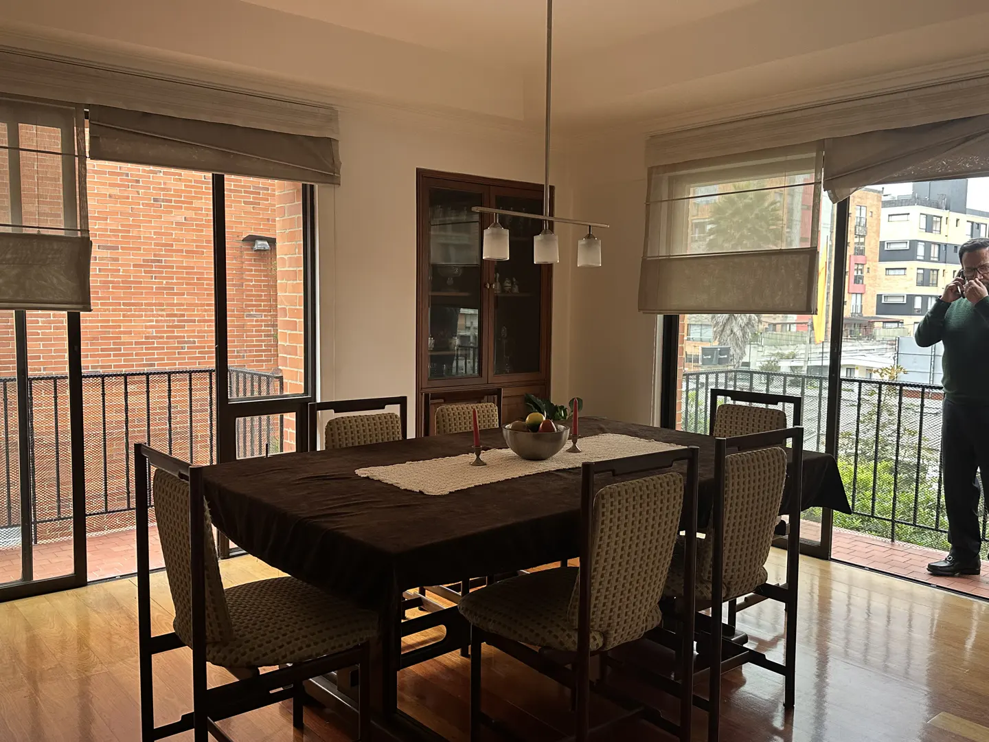 Dining room with a dark wood table and chairs, a man on the balcony, and a view of the city.