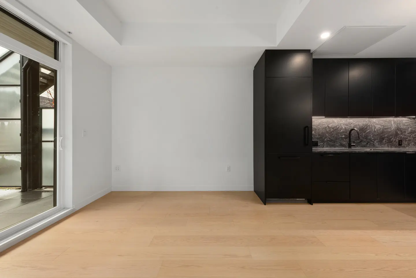 An empty room with light wood floors, white walls, and black kitchen cabinets. A sliding glass door is on the left.