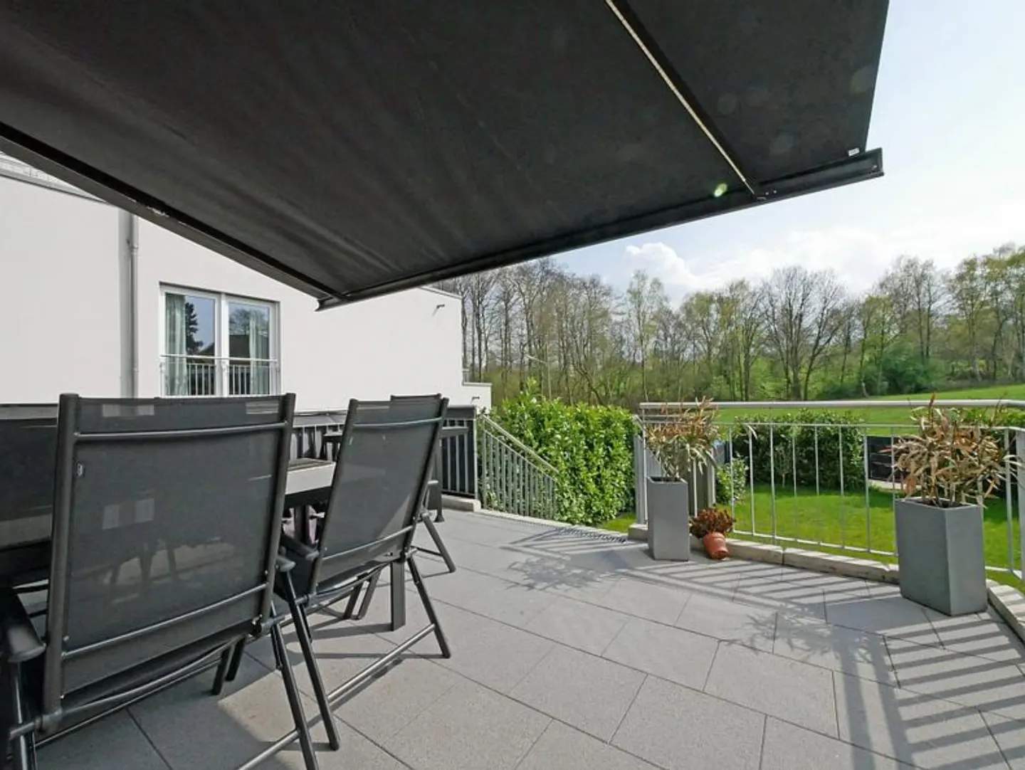 A patio with a black awning, gray chairs, and a table overlooks a green lawn and trees.