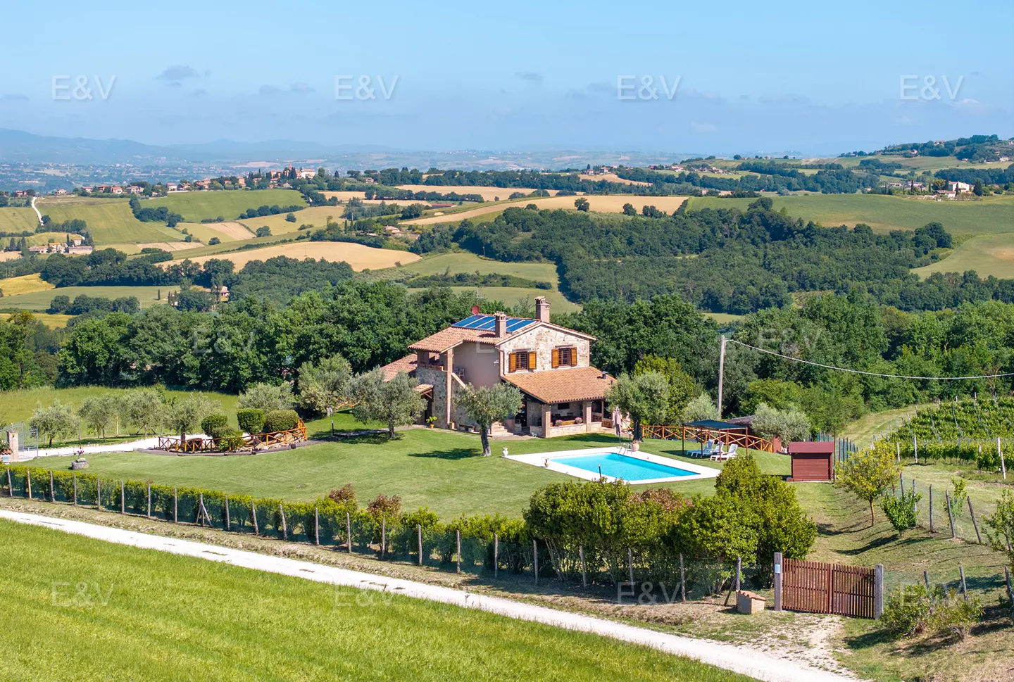 Tuscan villa with pool, stone facade, and red tile roof, set in a green, rolling landscape under a blue sky.