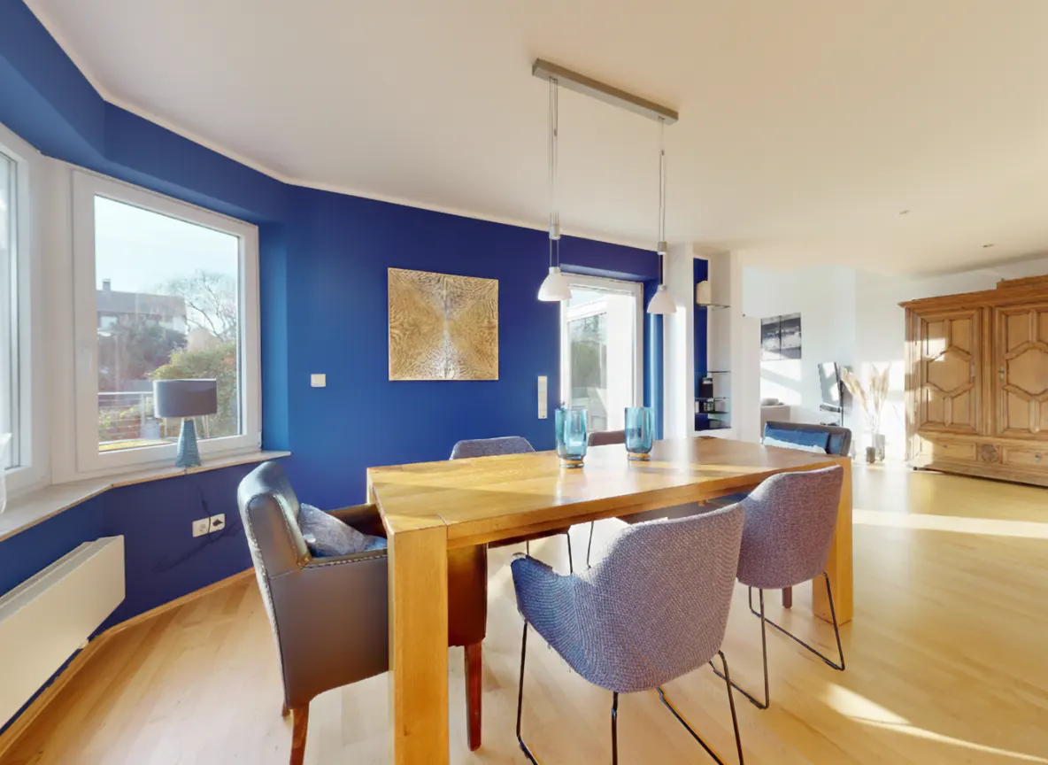 Bright dining room with blue walls, a wooden table, and upholstered chairs. A large window and artwork add to the ambiance.