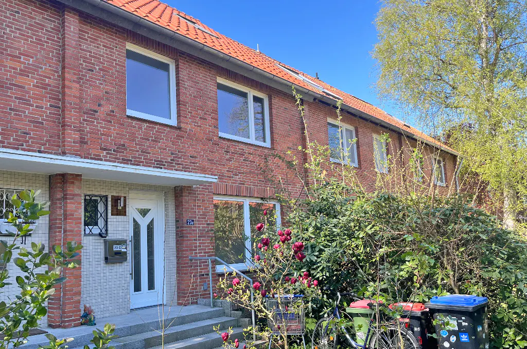 A red brick townhouse with a white door and windows, under a blue sky. Bushes and trees surround the building.