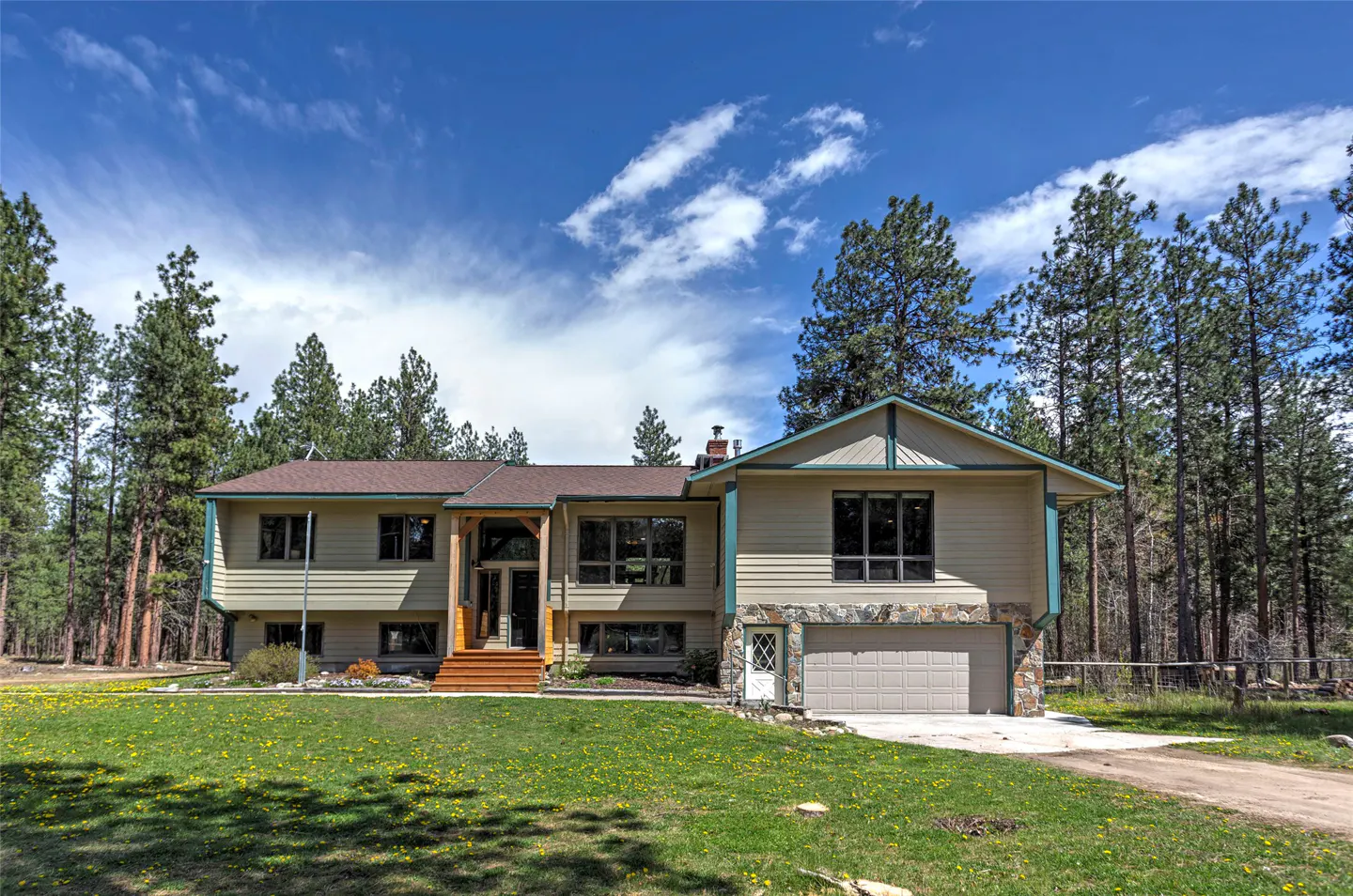 Two-story beige house with a brown roof, stone accents, and a green lawn with yellow flowers. Tall pine trees surround the property.