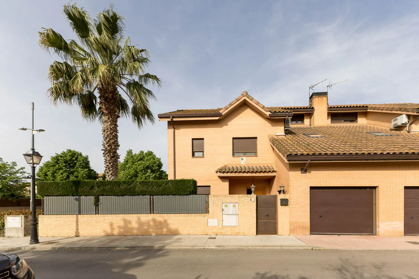 Two-story tan brick house with brown garage doors and tile roof. A tall palm tree stands to the left.