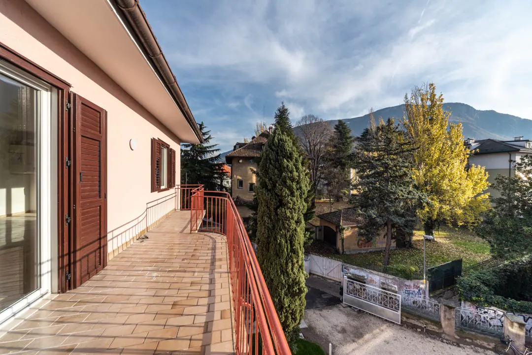 Balcony view of a peach-colored building with brown shutters and a red railing, overlooking trees and a mountain range.