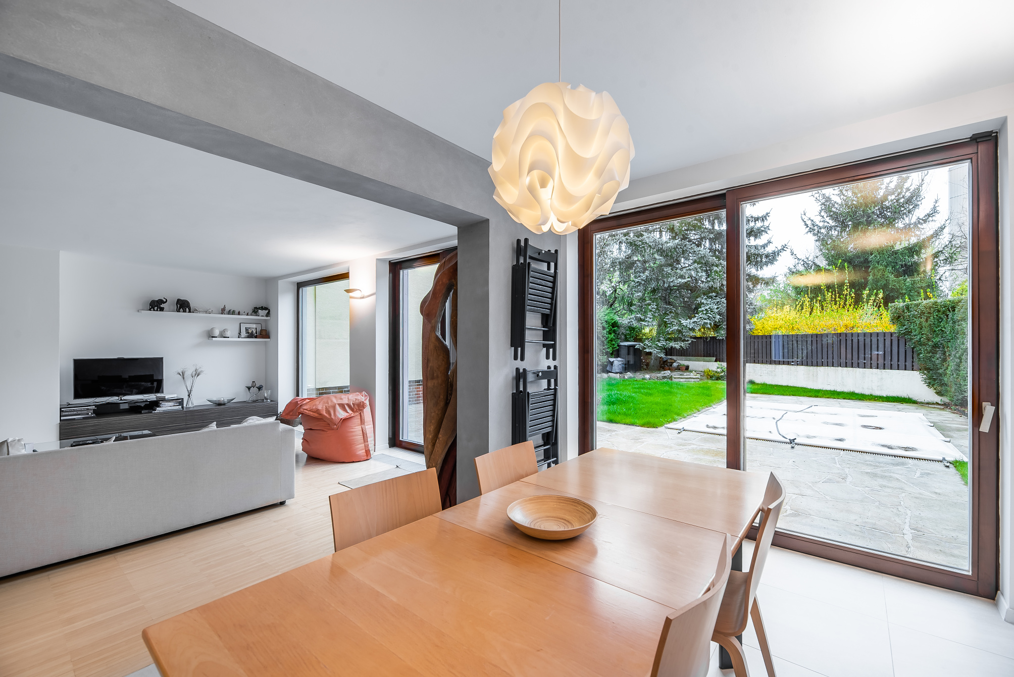 Bright, modern dining room with a light wood table, chairs, and a white pendant light. Sliding glass doors open to a green lawn and garden.