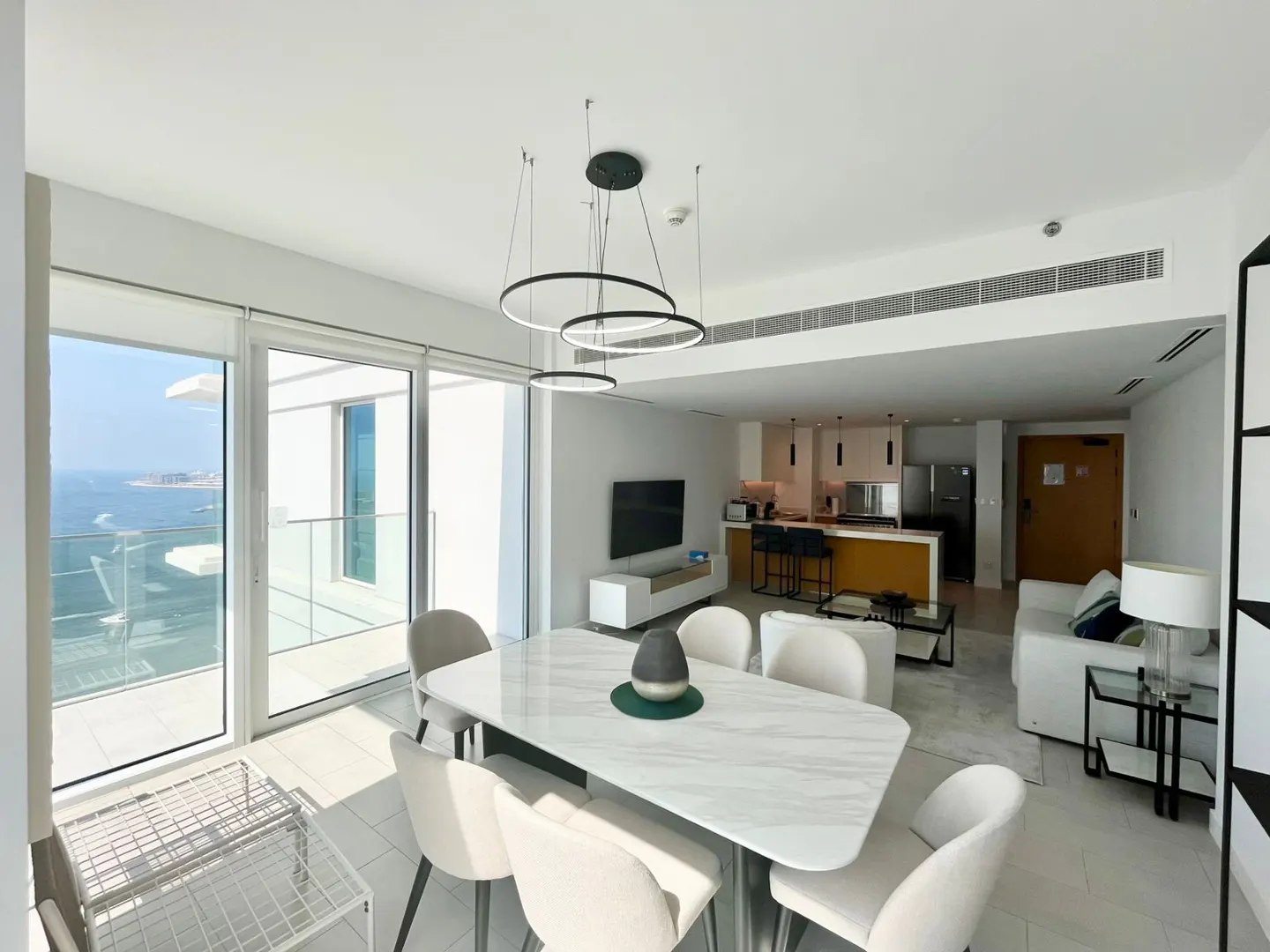 Bright, modern apartment interior with a marble dining table, white chairs, and a view of the ocean from a large window.