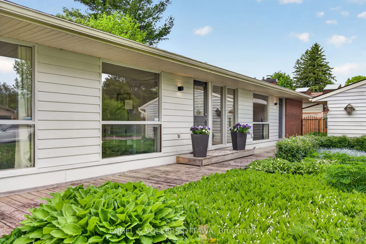 Exterior of a white, single-story home with large windows, a wooden walkway, and lush green landscaping.