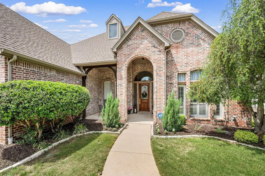 Brick home with arched entryway and wooden door. Green lawn and landscaping. Blue sky with clouds.