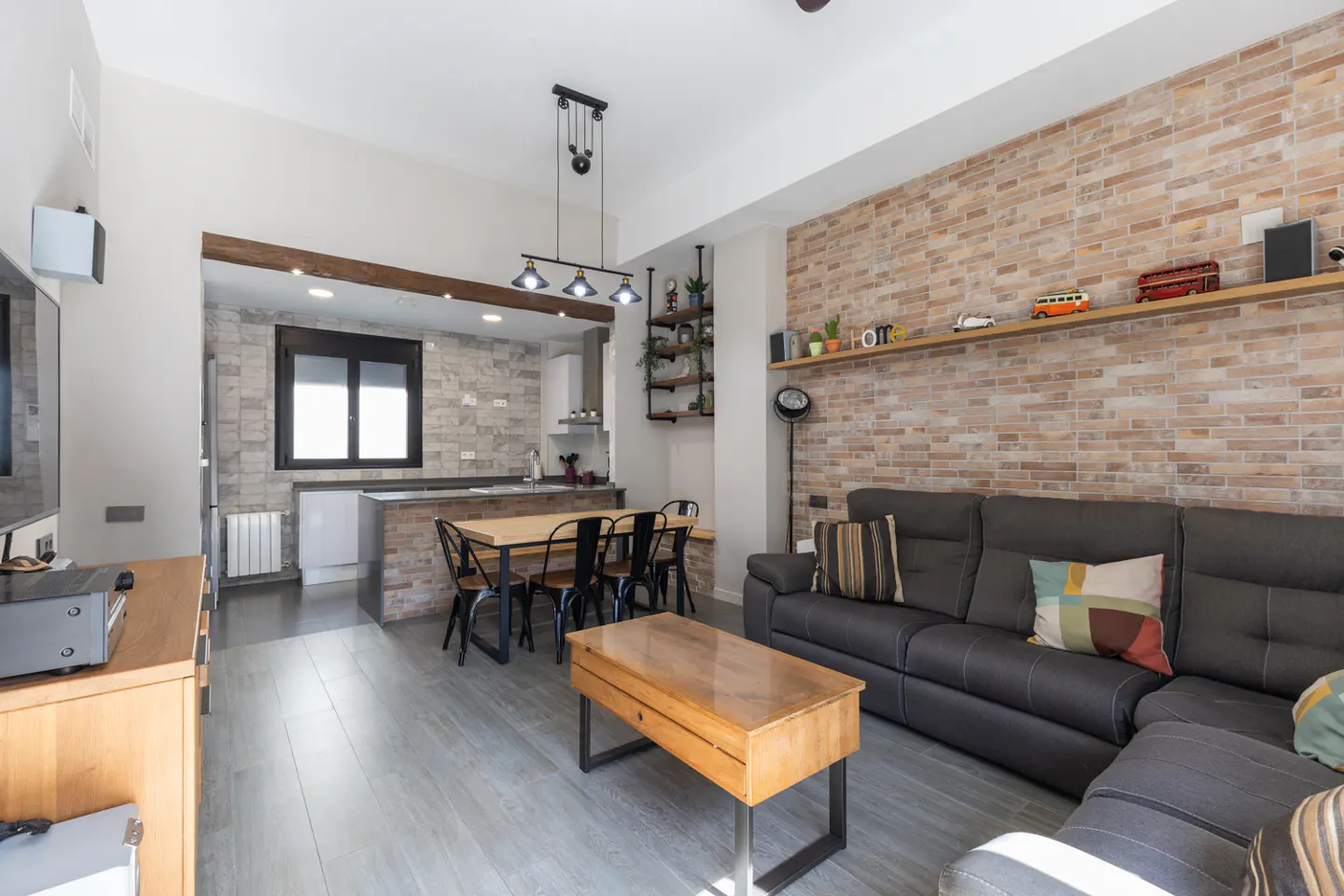 Open-concept living space with gray sofa, wood coffee table, and dining table. Brick accent wall and gray wood floors.
