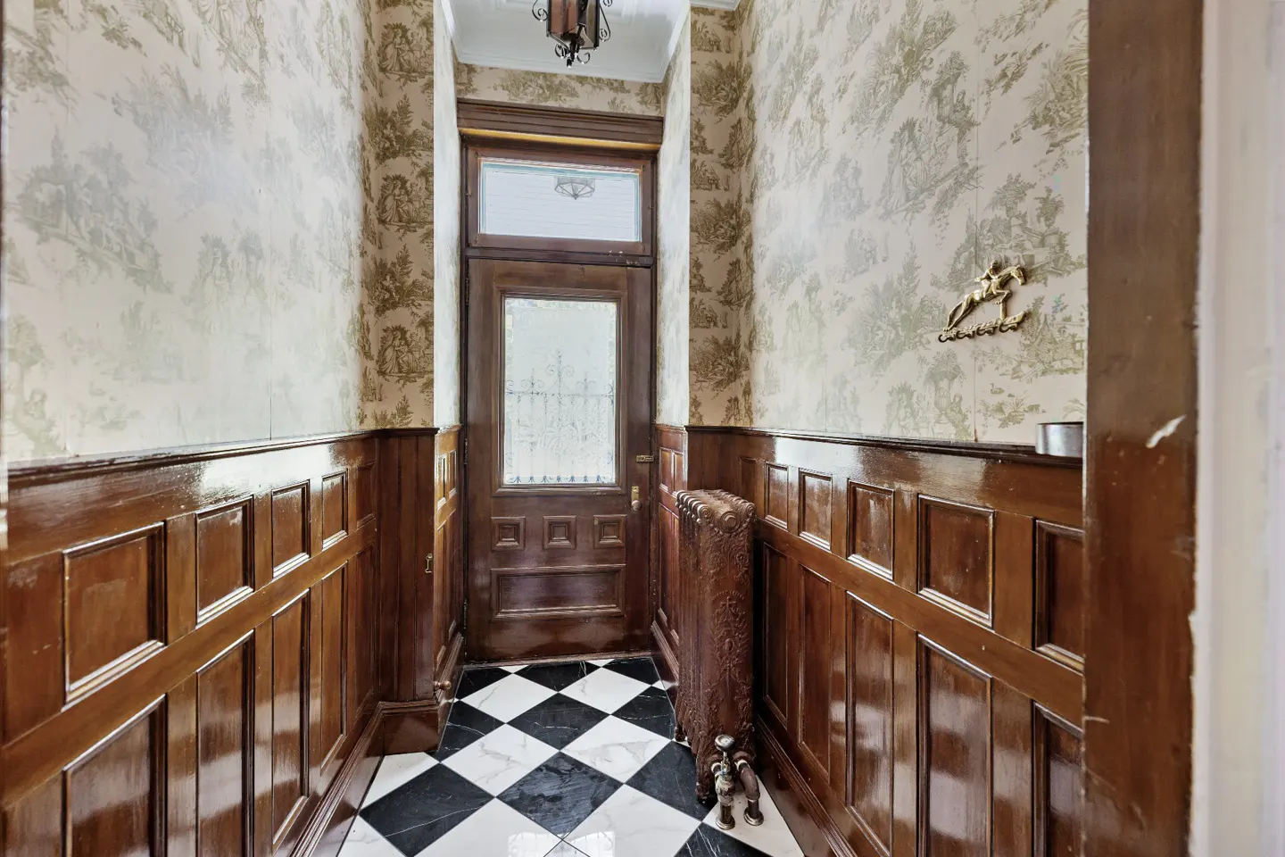 Hallway with black and white checkered floor, wood paneled walls, and patterned wallpaper. A wooden door with a window is at the end.