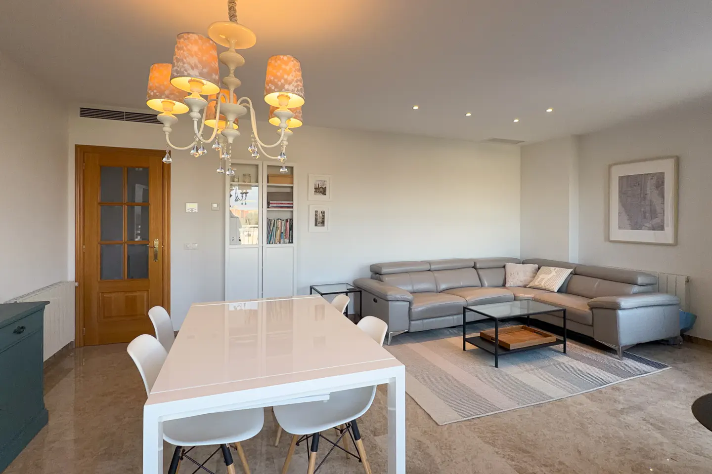 Bright living room with a white table, chairs, and a gray leather sectional sofa. A decorative chandelier hangs above the table.