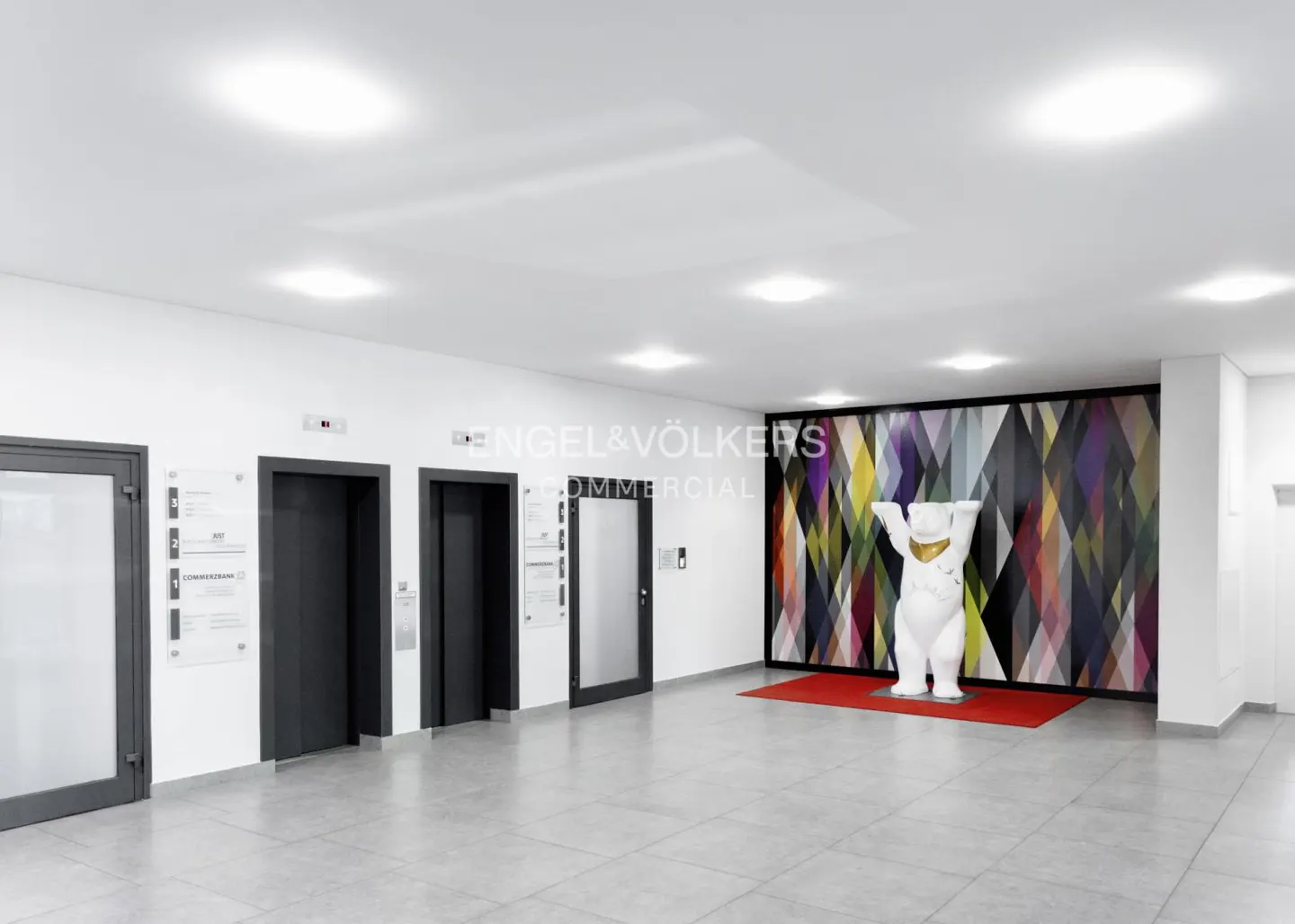 Bright lobby with gray tile floor, white walls, and recessed lighting. Two elevators and a glass door are on the left. A white bear statue stands on a red rug in front of a colorful geometric wall.