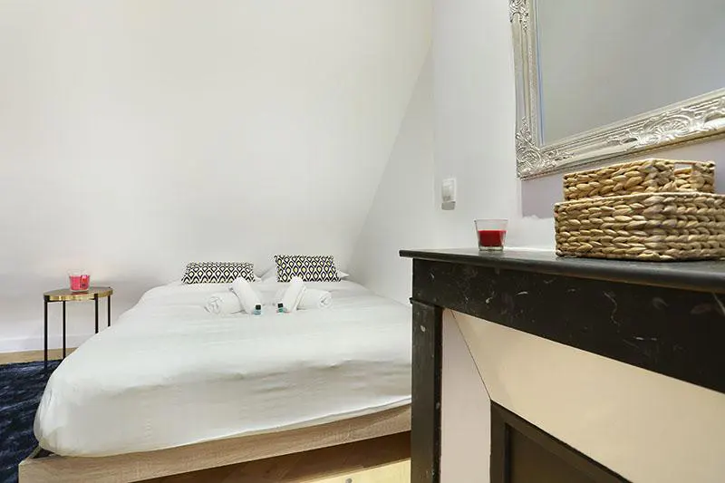 A bedroom with a white bed, patterned pillows, a small side table, and a black fireplace mantel with a mirror above it.