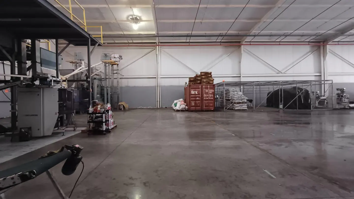 A wide shot of a large, empty warehouse with machinery, containers, and stacked goods.