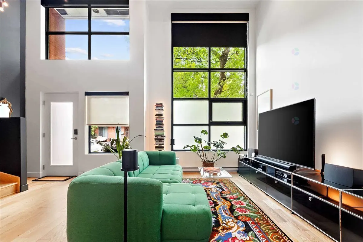 Bright living room with a green sectional sofa, large windows, and a black media console with a TV. A colorful rug adds warmth to the light wood floors.