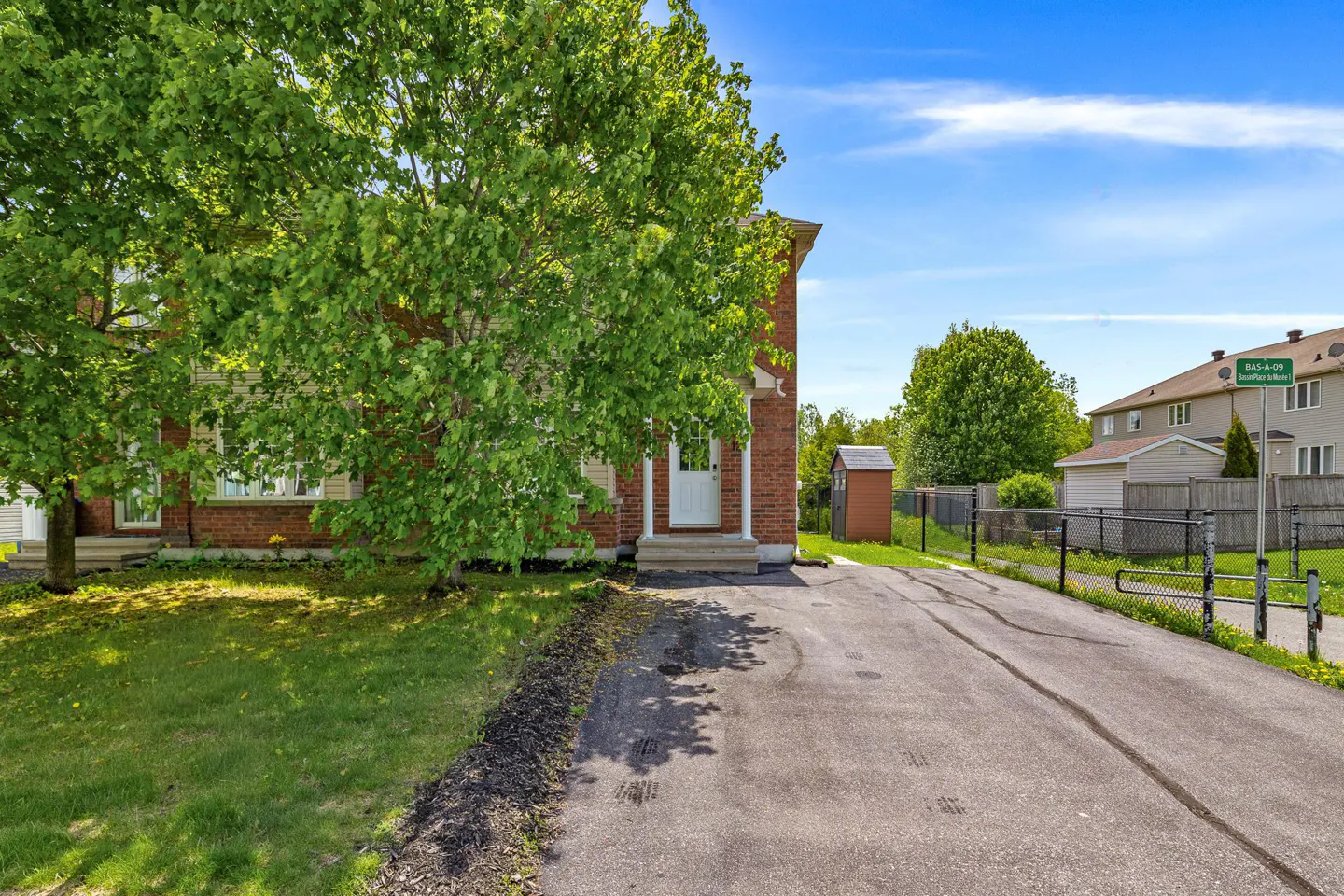 A brick house with a white door, a large tree in the front yard, and an asphalt driveway under a bright blue sky.