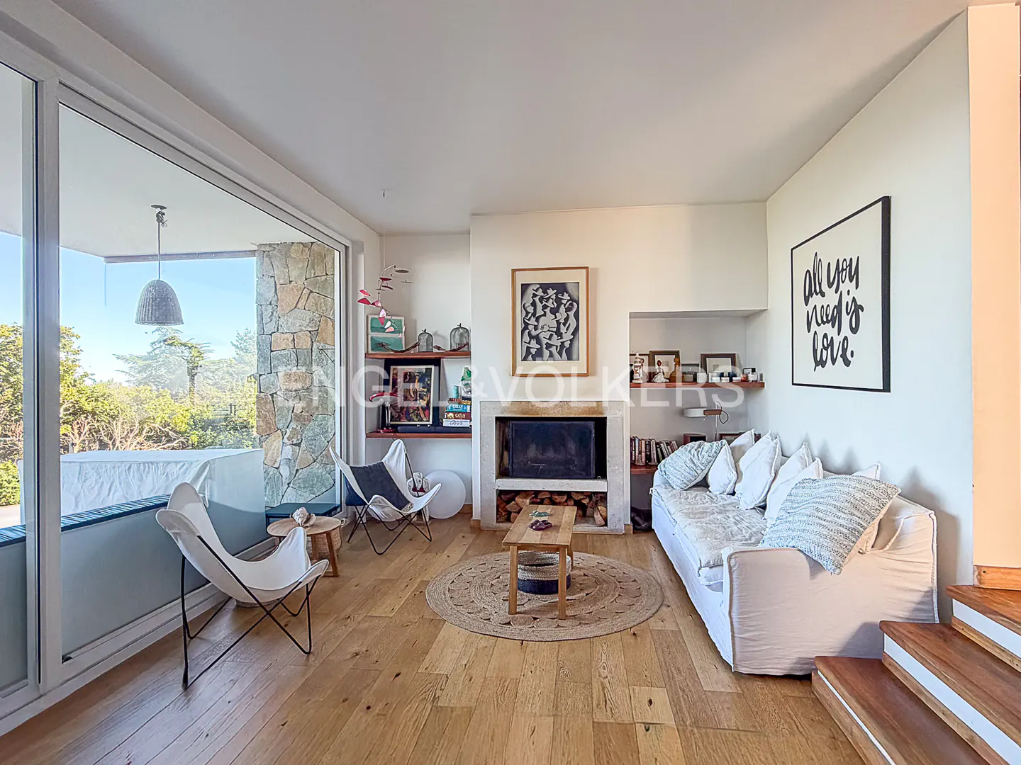 Bright living room with wood floors, white sofa, fireplace, and large windows overlooking greenery. Butterfly chairs and a round rug add to the cozy feel.