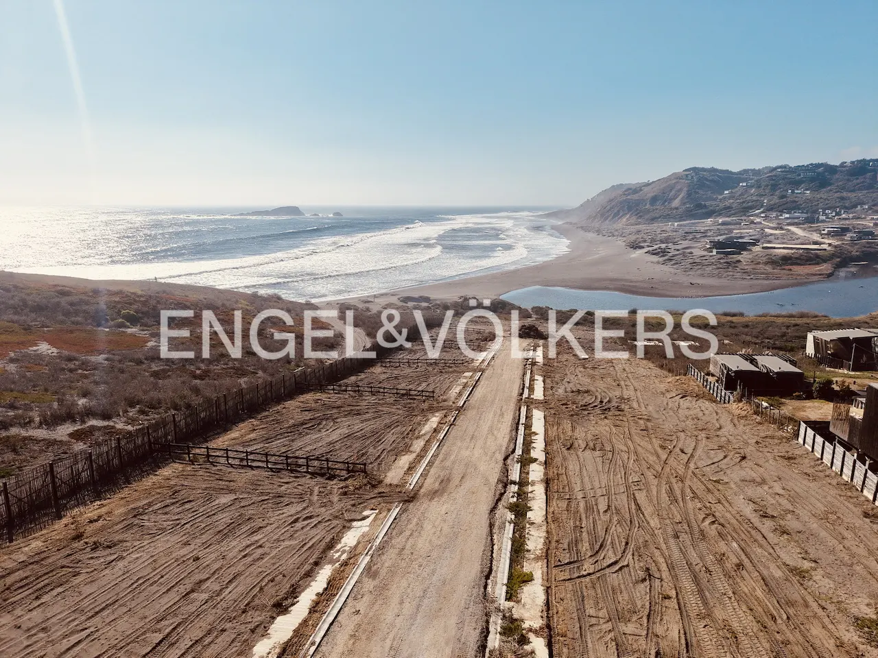 Aerial view of vacant land plots near a beach with ocean waves under a clear blue sky. Engel & Völkers logo overlay.