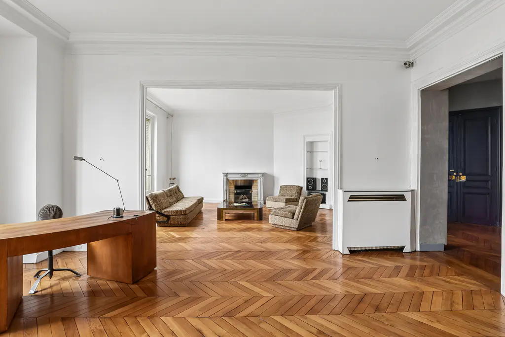 Bright, open-plan apartment with herringbone wood floors. A desk sits in the foreground, with a living room visible through an archway.