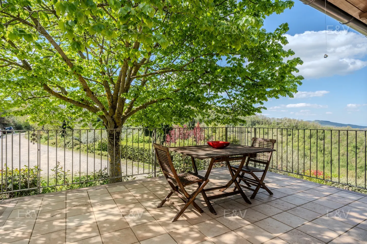 Outdoor patio with a wooden table and chairs under a large tree, overlooking a green landscape on a sunny day.