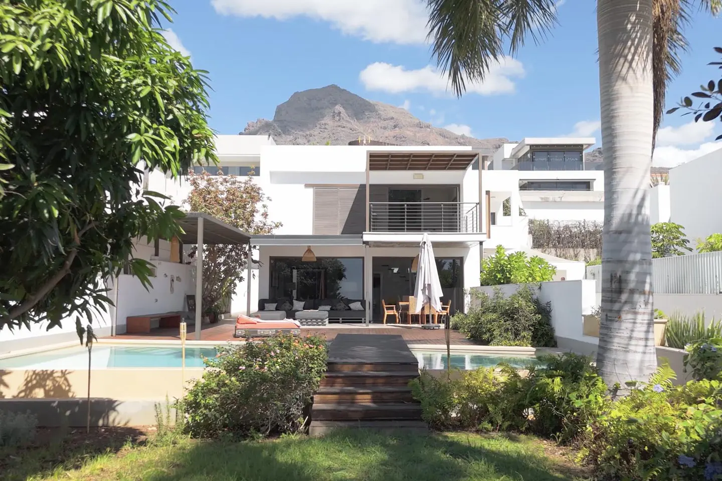 Modern white house with a pool, wooden deck, and mountain backdrop under a blue sky with palm trees and green foliage.