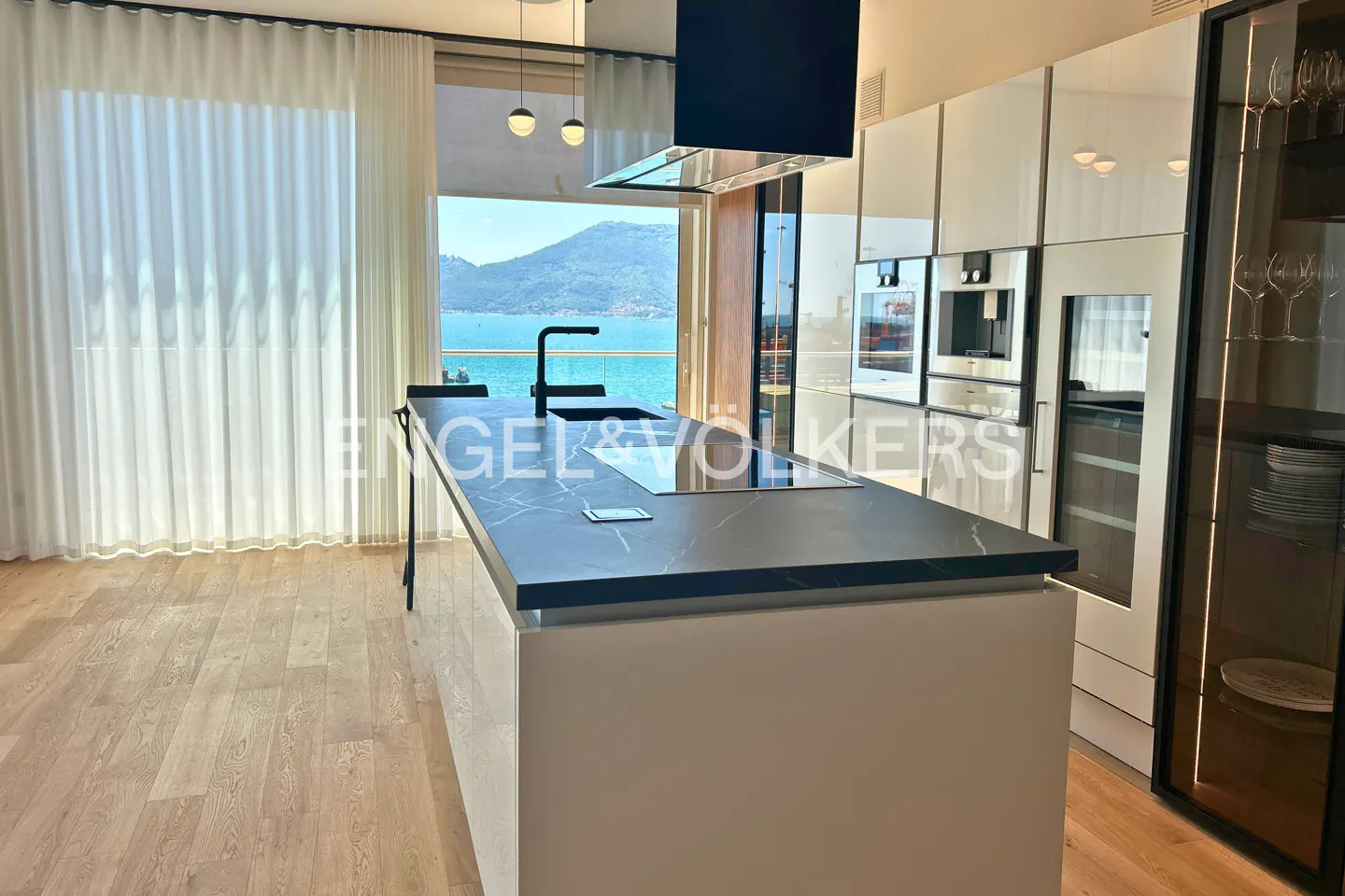 Modern kitchen with a black marble island, white cabinets, and a scenic view of the blue sea and mountains.