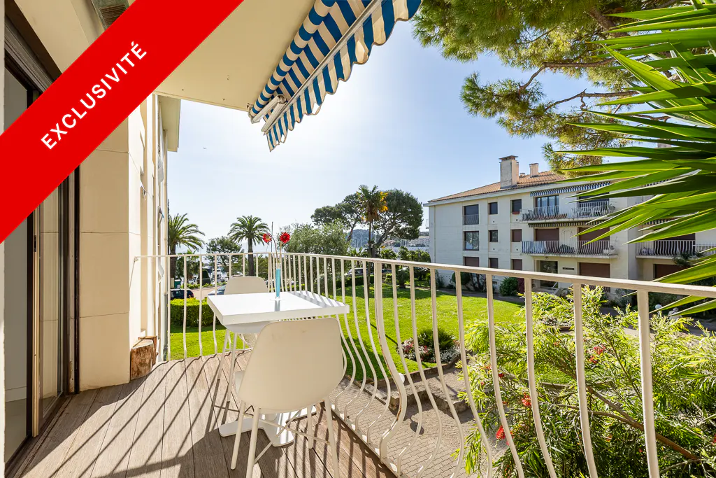 Balcony view with white table and chairs, blue and white awning, and a red "Exclusivite" banner. Green lawn and building in the background.
