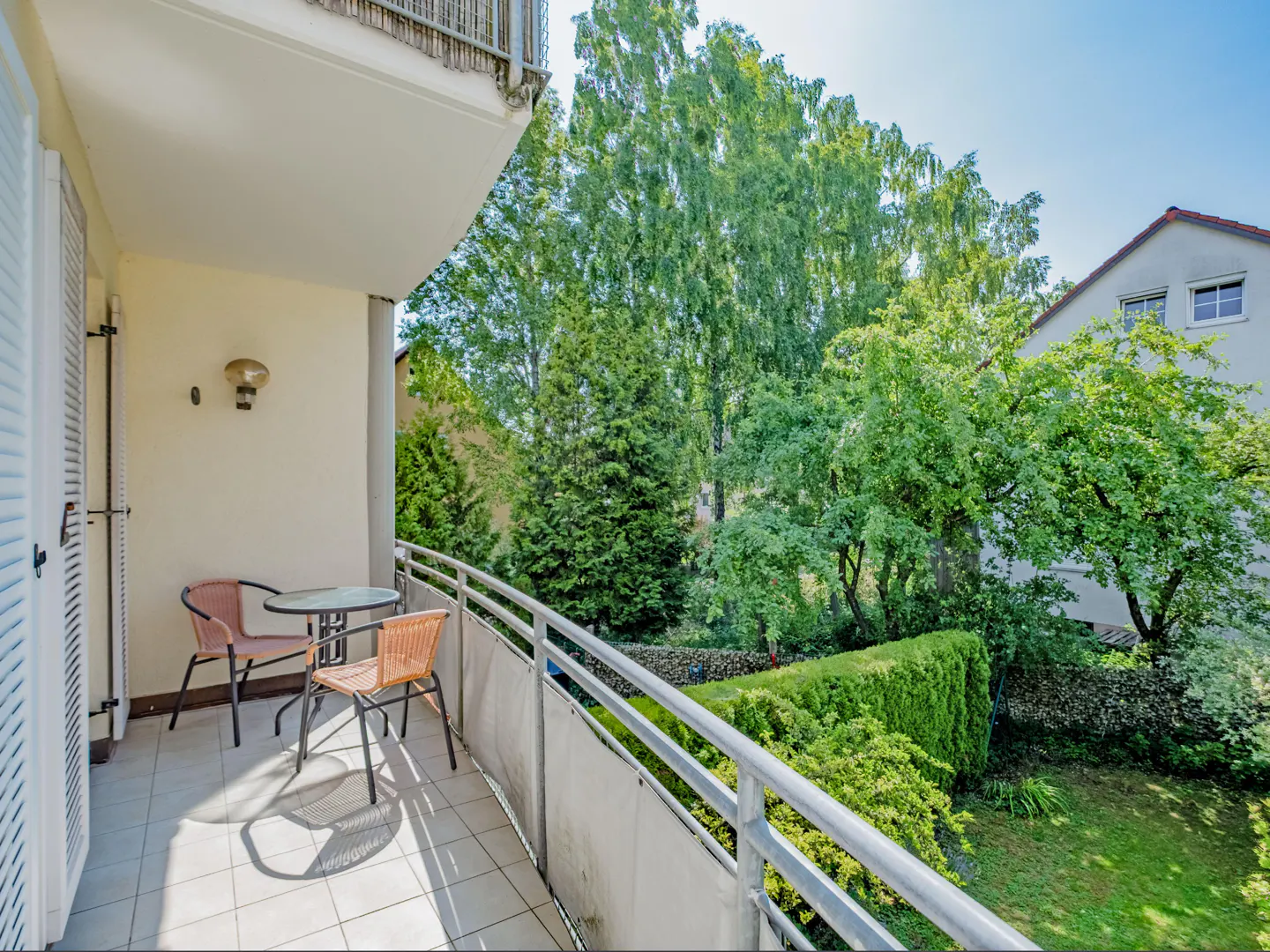 Balcony with a table and two chairs overlooking a lush green garden on a sunny day.